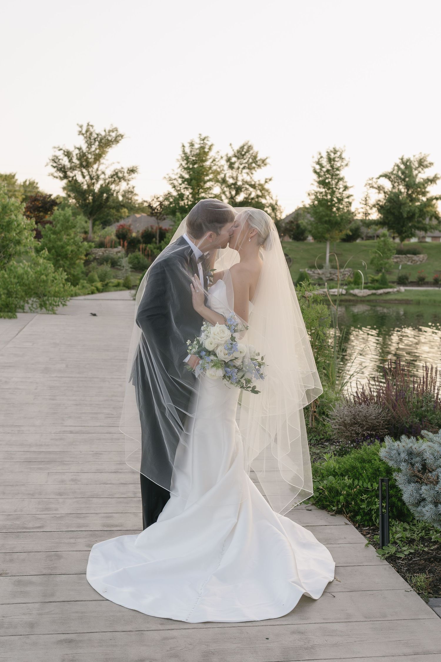 Bride and groom kissing under veil on wooden dock beside pond, surrounded by lush greenery at outdoor wedding.