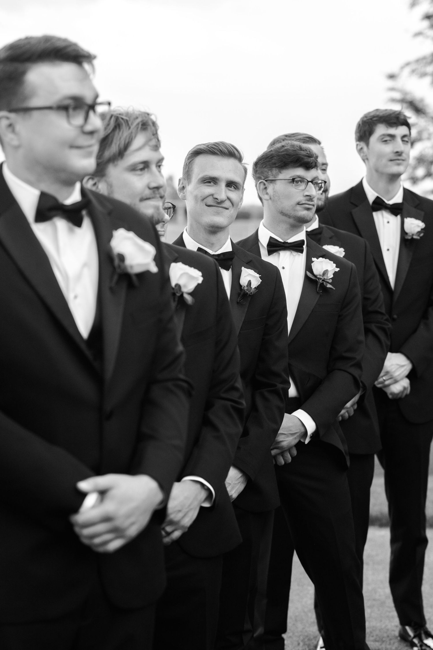 A black and white photo of groomsmen in matching tuxedos with white boutonnieres lined up at a formal event.