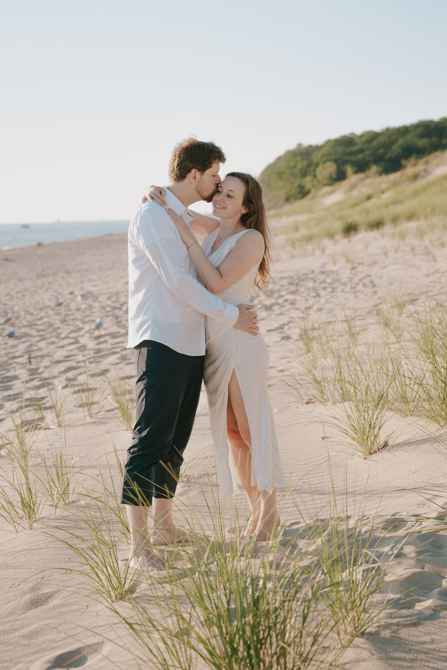 A romantic couple embraces on a sandy beach with dunes and ocean in the background.