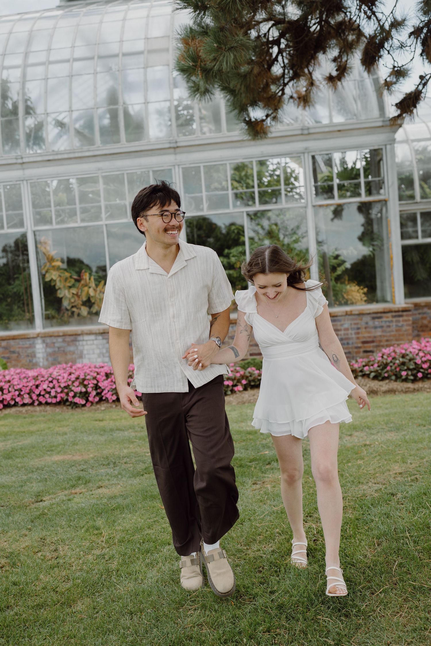 A couple takes playful steps together on the lawn in front of a conservatory building.