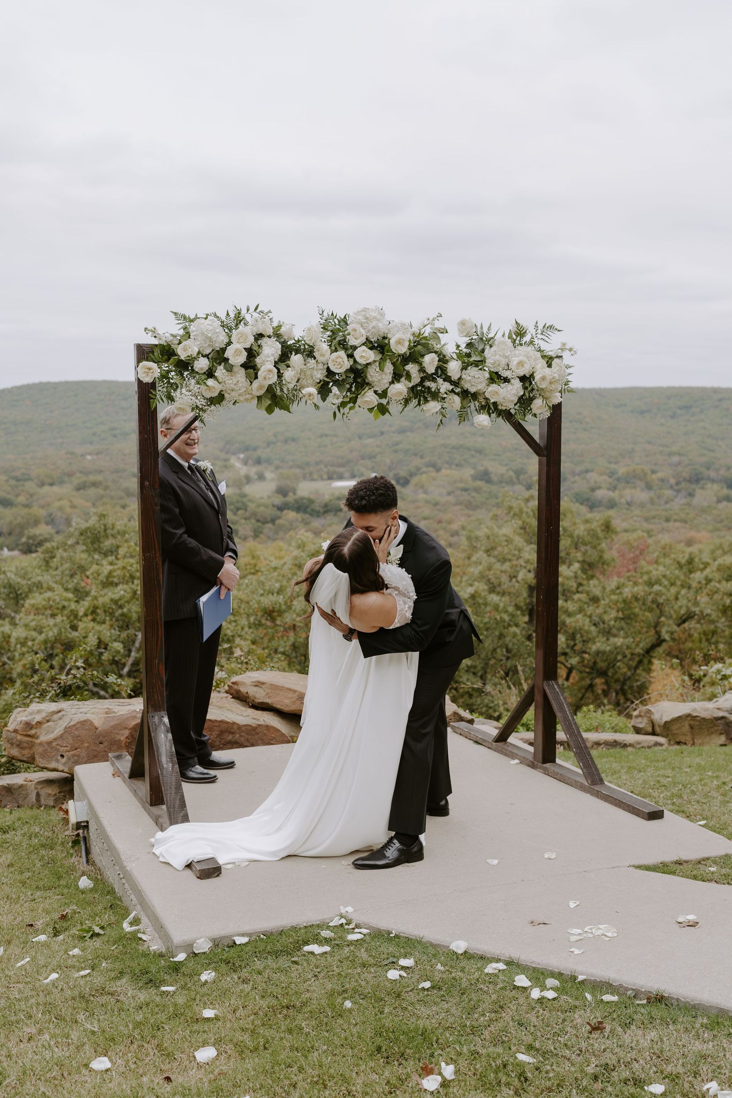 Outdoor wedding ceremony with wooden arch and white floral decorations against rolling hills.