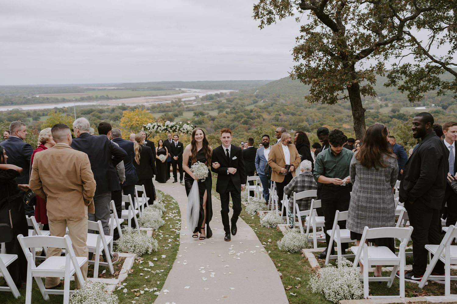 Wedding guests stand beside white chairs on a scenic hillside path during the ceremony recessional.