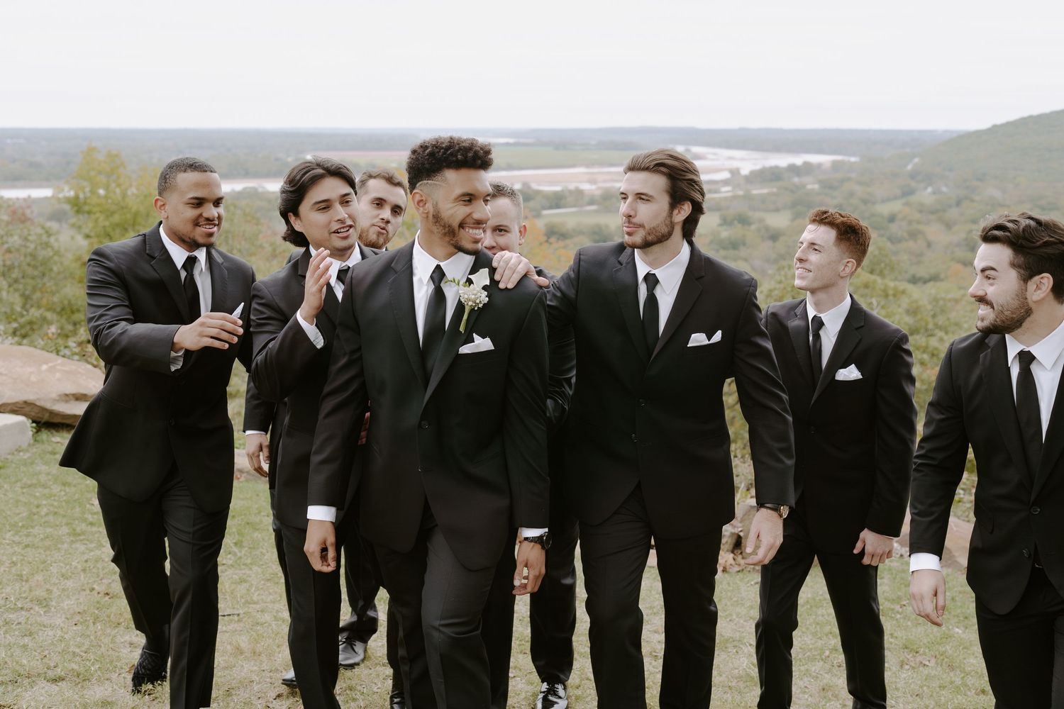 Groomsmen in black tuxedos share jokes and laughter during an outdoor photo session.
