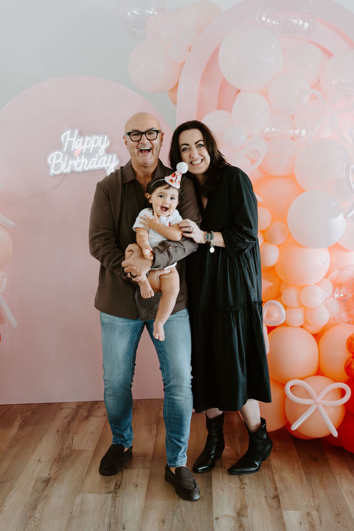 A family portrait with a baby in front of a 'Happy Birthday' balloon display.