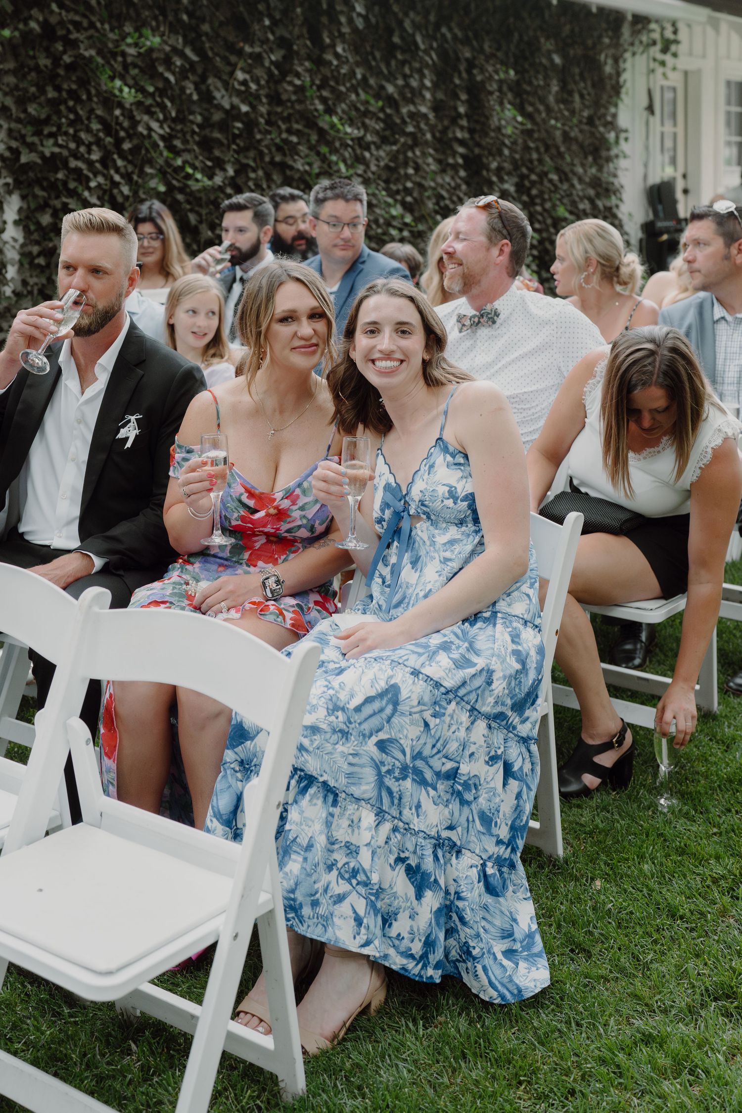 Wedding guests in blue floral dresses sit on white chairs on a grassy lawn during an outdoor ceremony.
