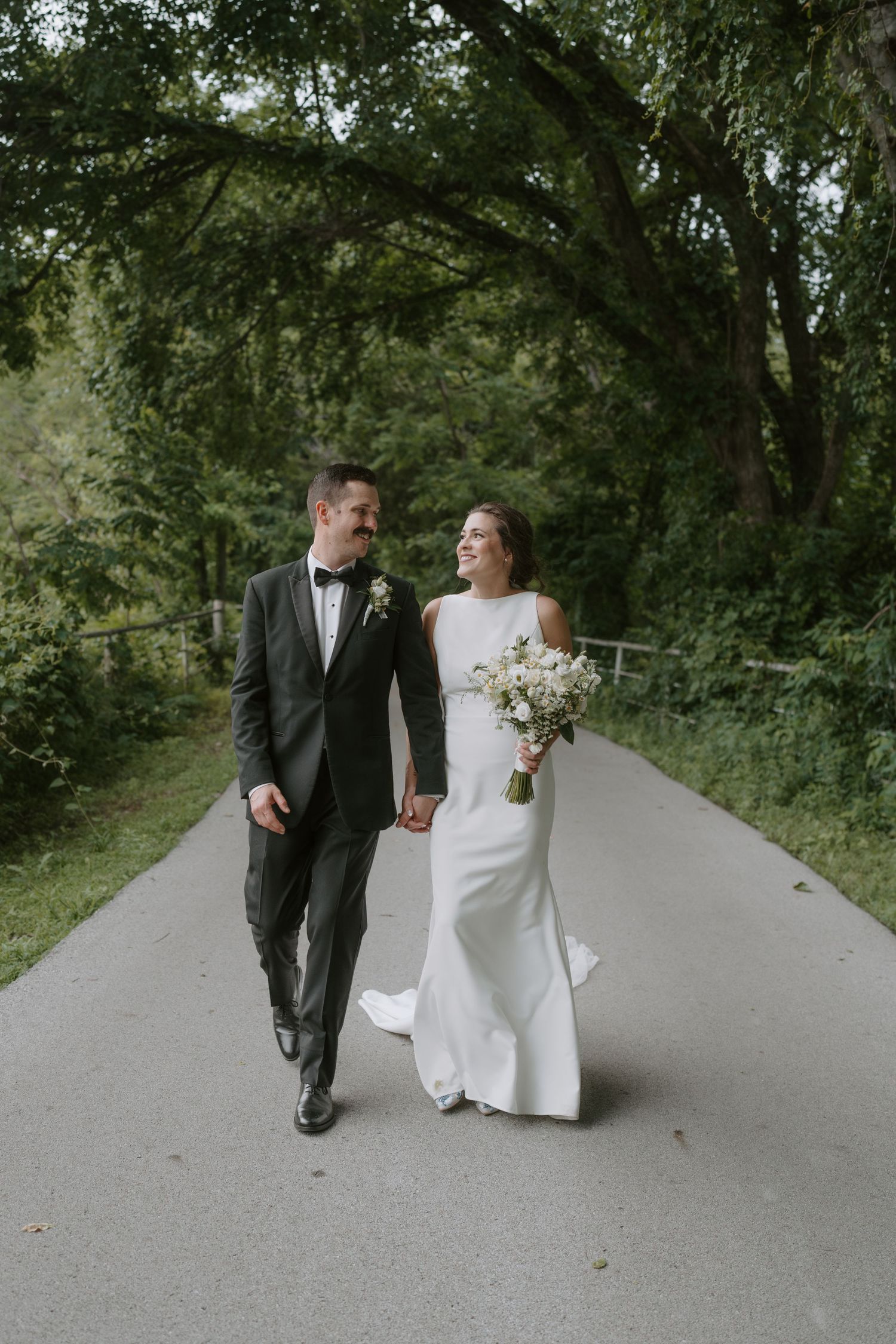 Newlyweds walk hand in hand down a paved pathway with the bride holding a white floral bouquet.
