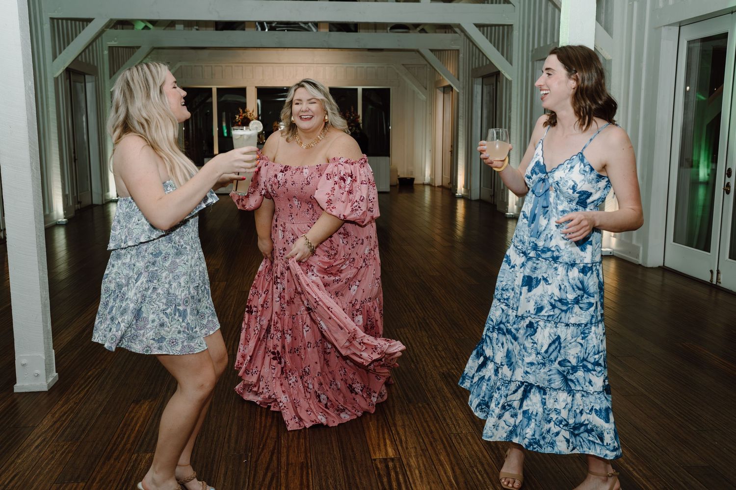 Guests in colorful floral dresses enjoy dancing at a wedding reception in a bright indoor venue.