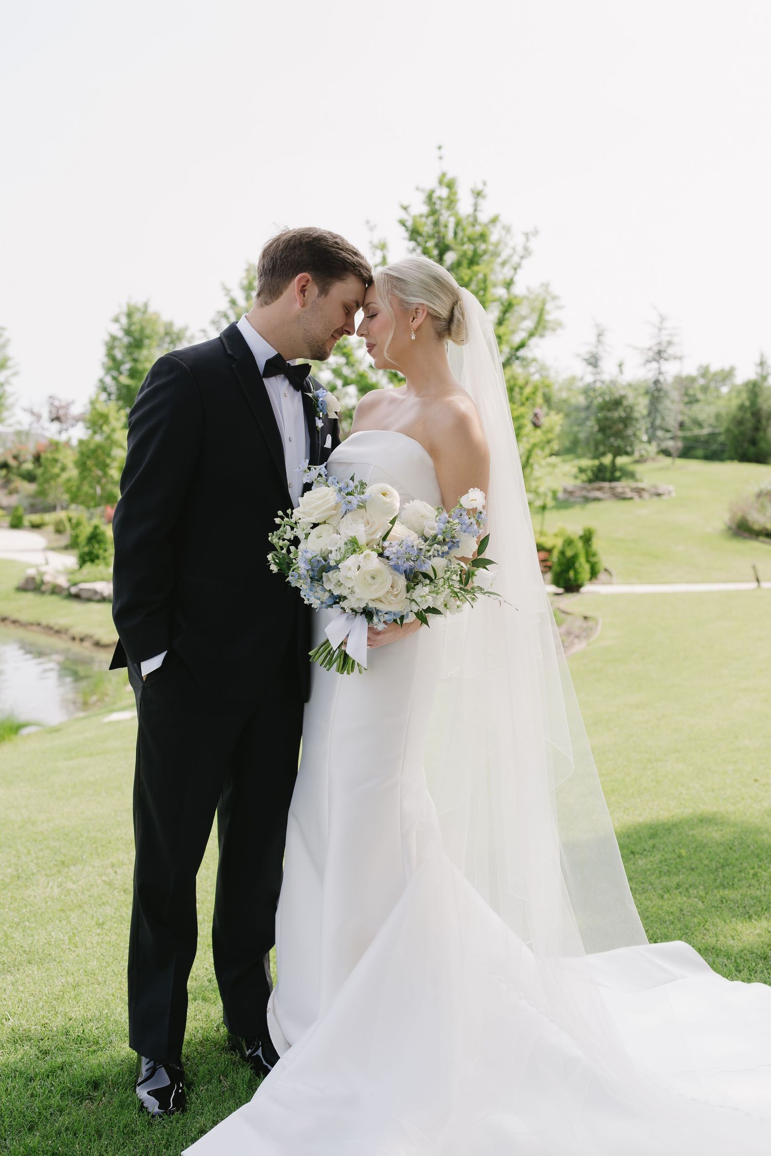 Bride and groom in formal wedding attire share a romantic forehead touch outdoors on lush green grounds.