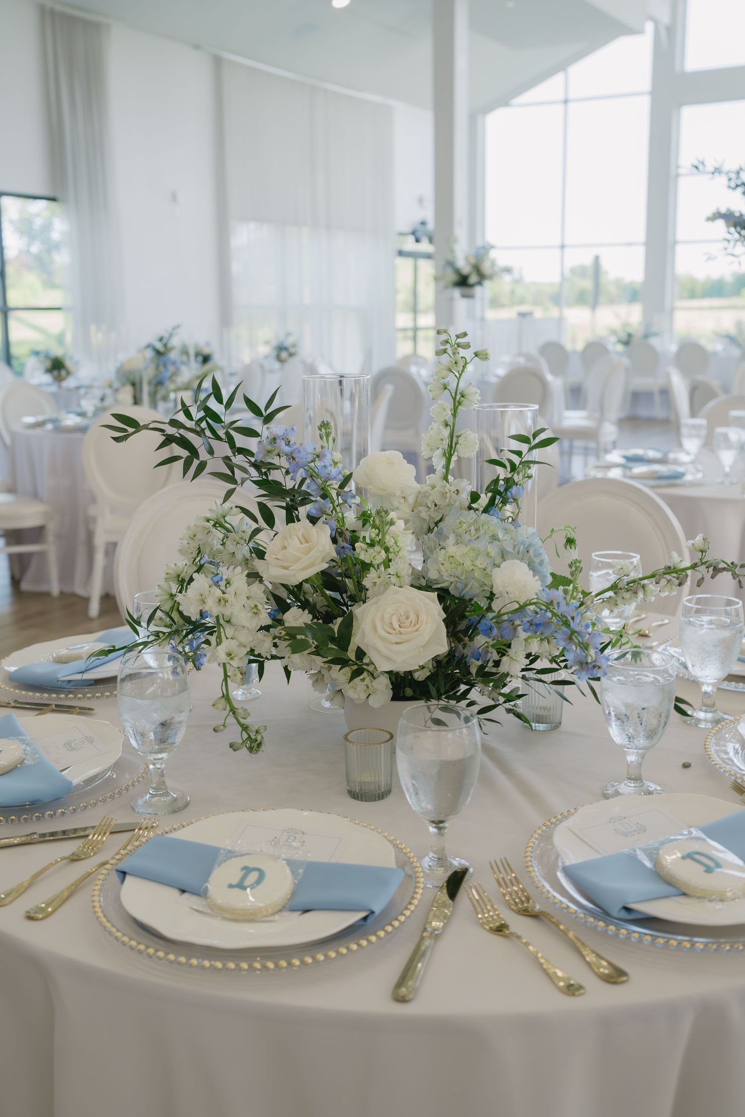 Elegant wedding reception table with white and blue floral centerpiece, gold cutlery, charger plates, and light blue napkins.