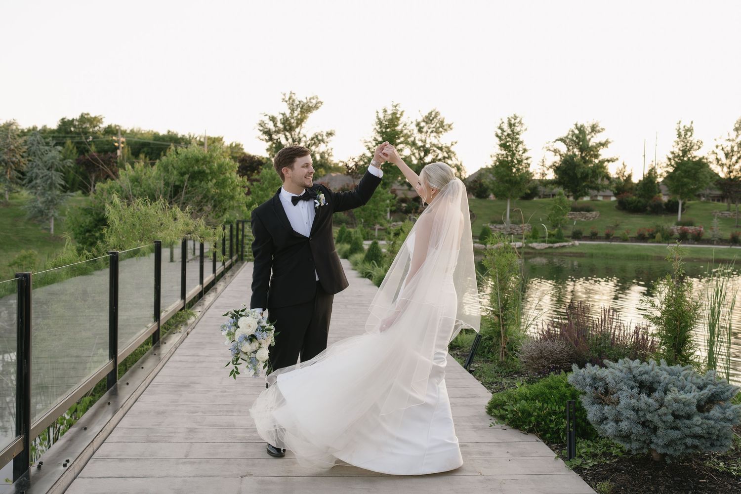 Bride and groom dancing on wooden dock by pond, groom spinning bride in white gown and veil at outdoor wedding.