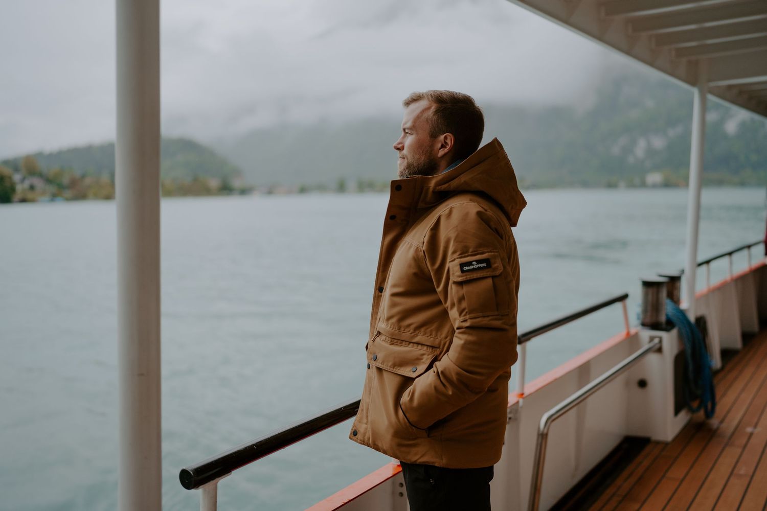 Person in brown coat standing on boat deck overlooking scenic mountain lake.