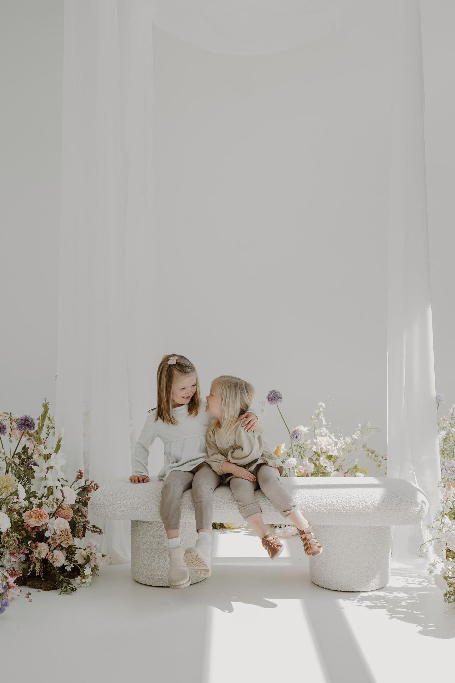 Two young children in white outfits sit together on a bench surrounded by floral arrangements in a bright studio setting.