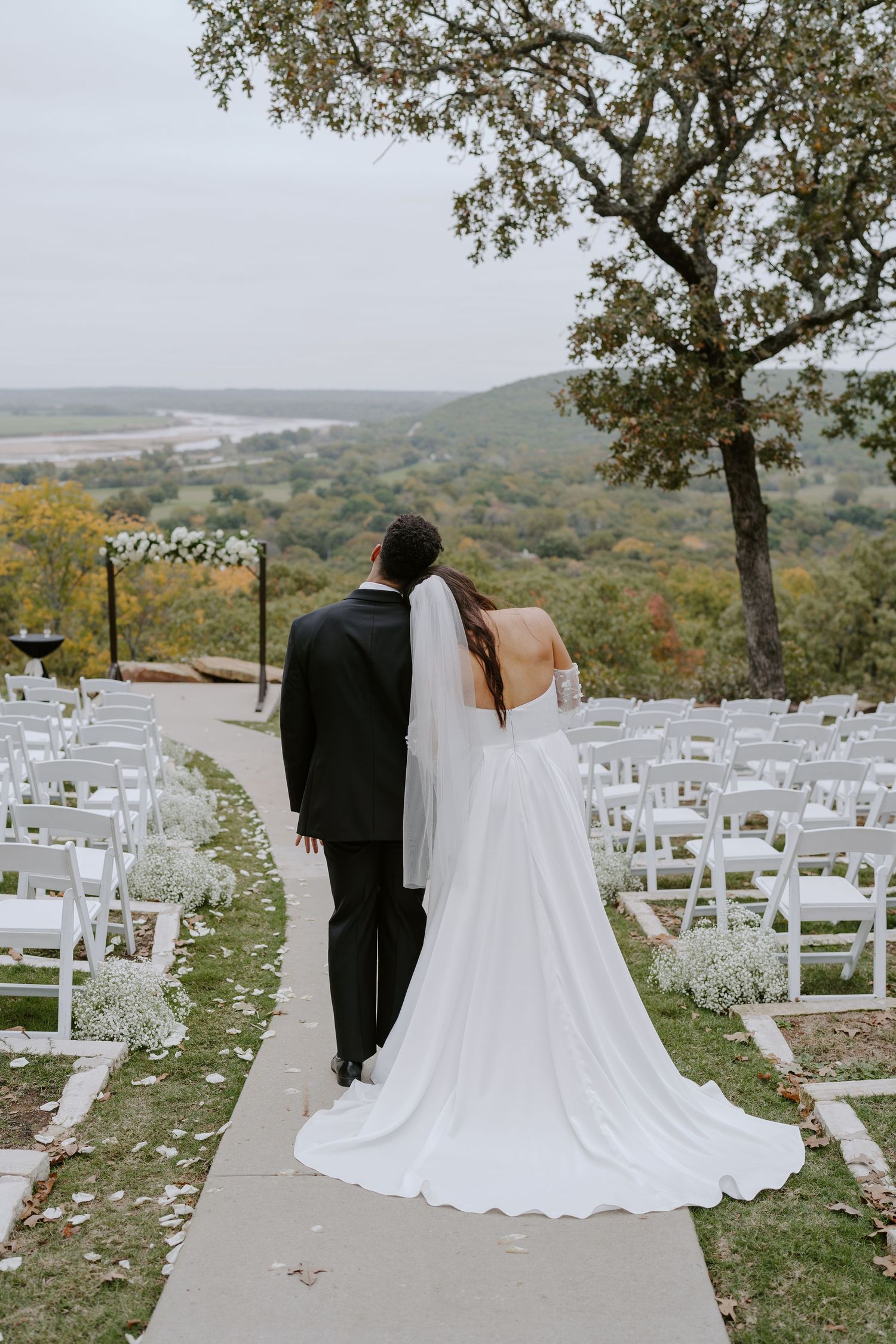 An elegant outdoor wedding venue with white chairs and fall foliage in the background.