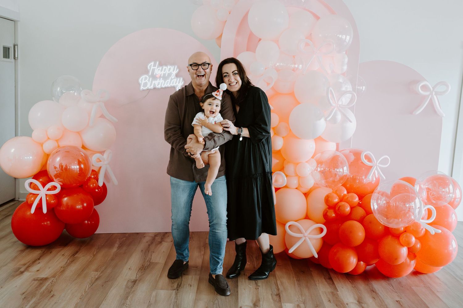 A couple holds a baby while standing in front of a festive balloon arch display.