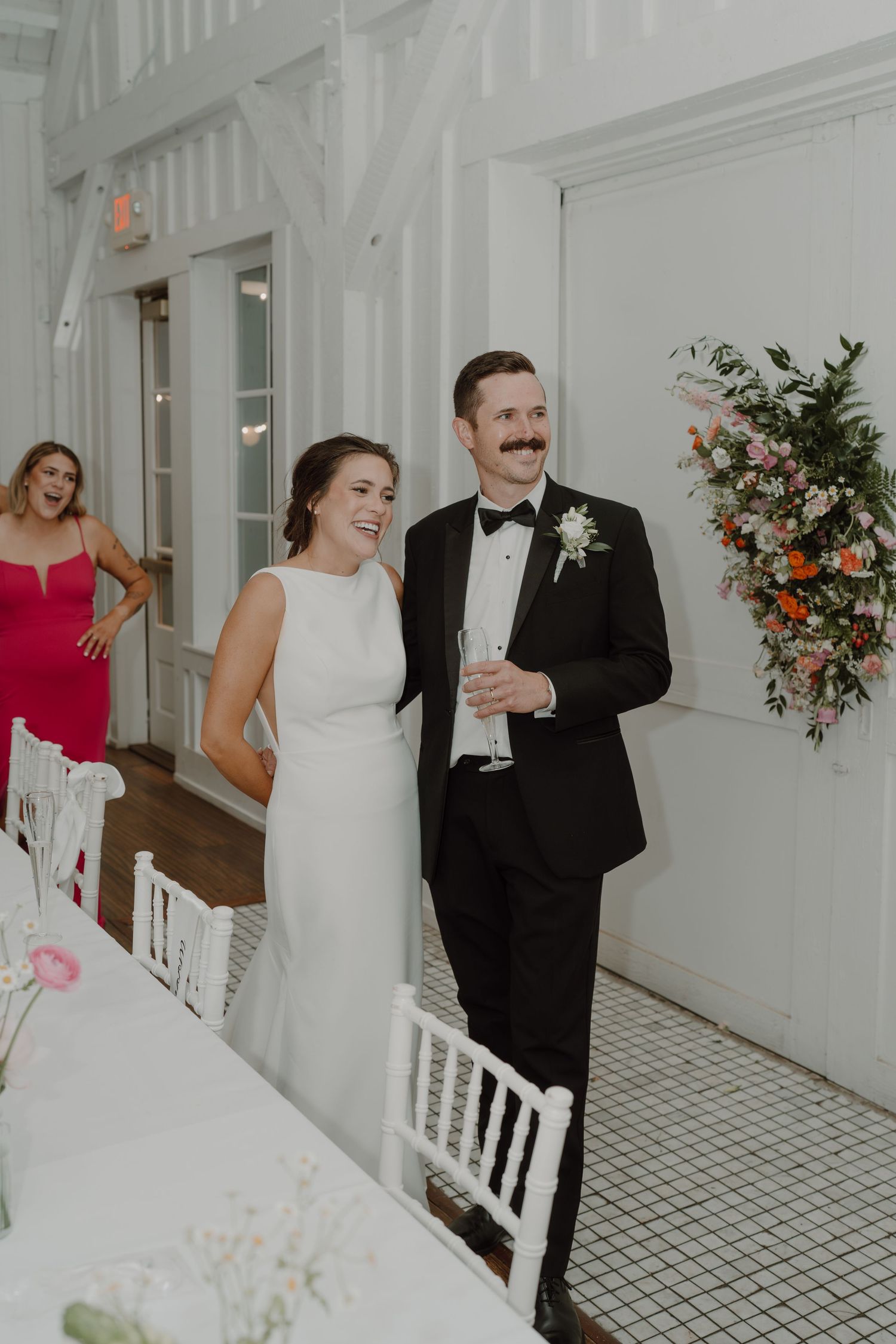 Wedding party members share joyful moments in an elegant white venue with floral decor.