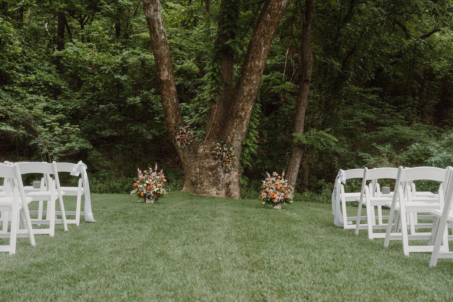 White ceremony chairs set up on a lush green lawn surrounded by mature trees.