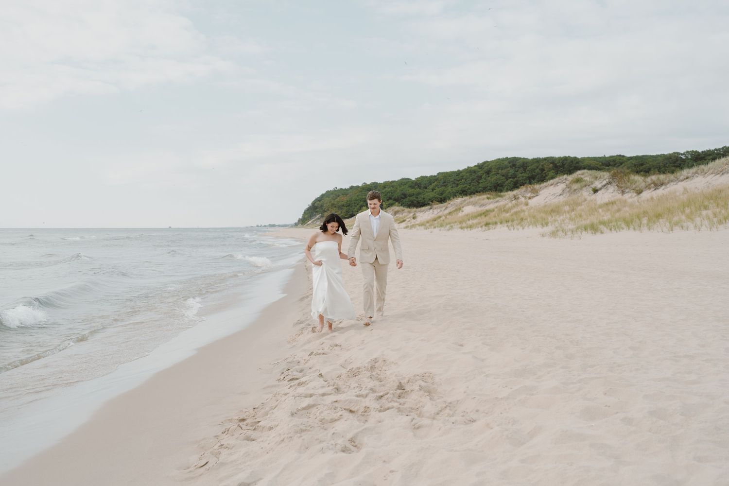 A couple walks along a pristine beach with sand dunes and ocean waves in the background.