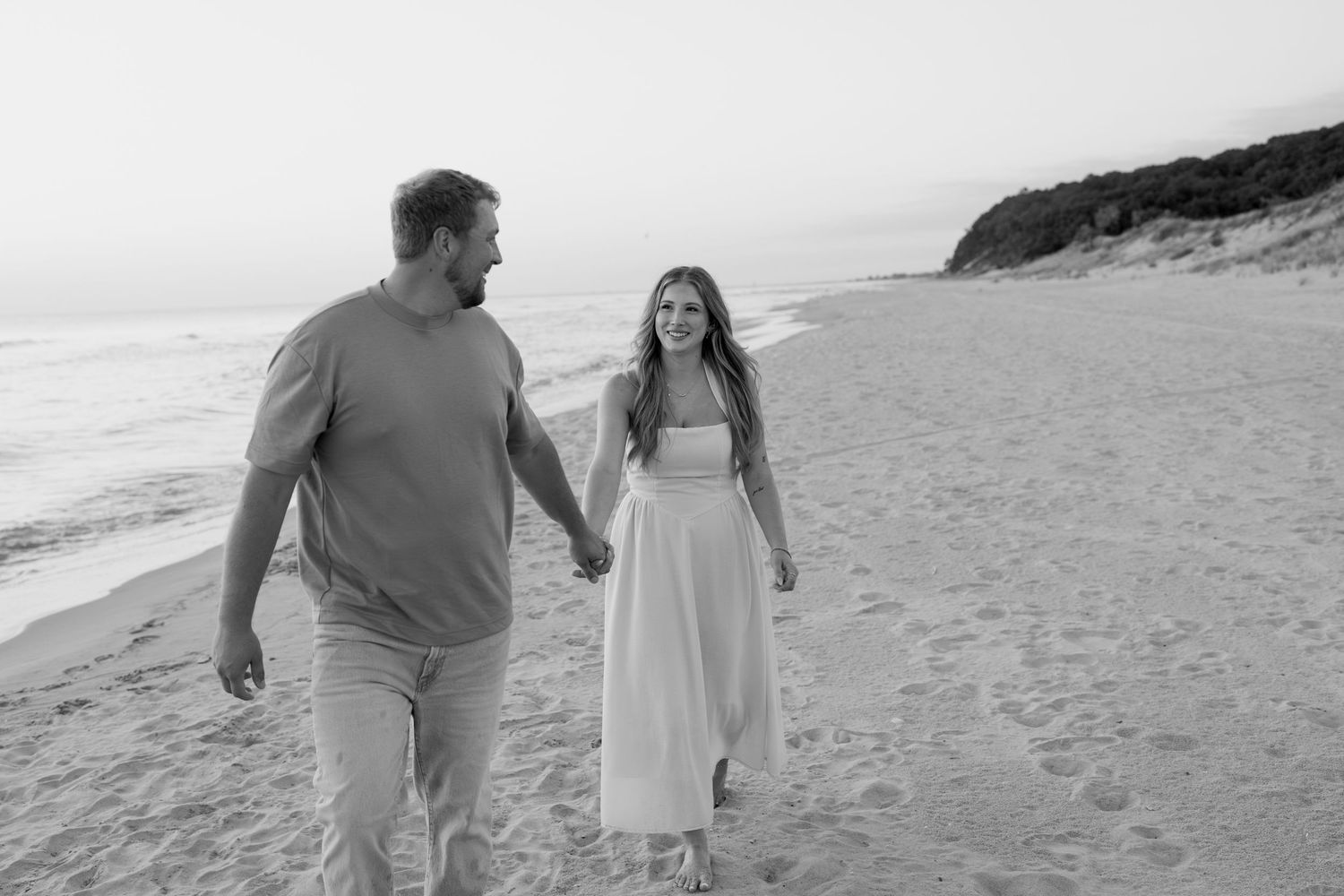 A couple strolls together on a sandy beach with cliffs and ocean waves visible in black and white.