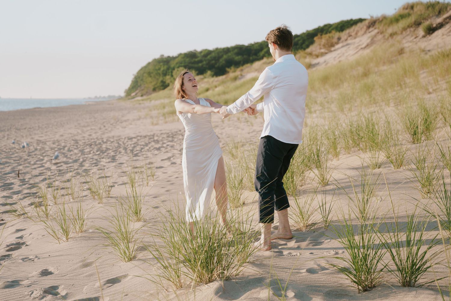 A playful moment between a couple dancing on the beach with coastal vegetation nearby.