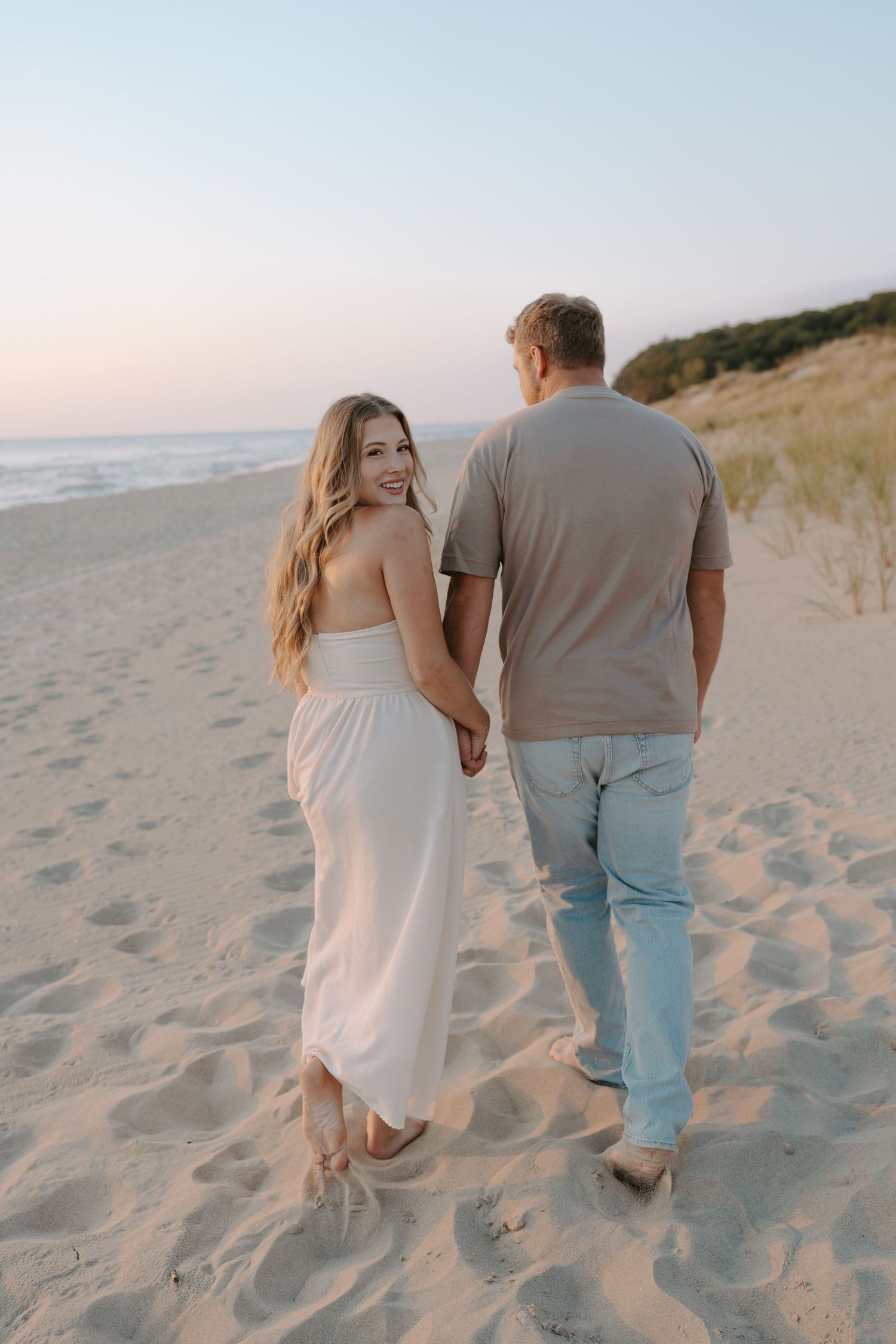 Couple strolling together on beach with dunes and ocean in background at dusk.