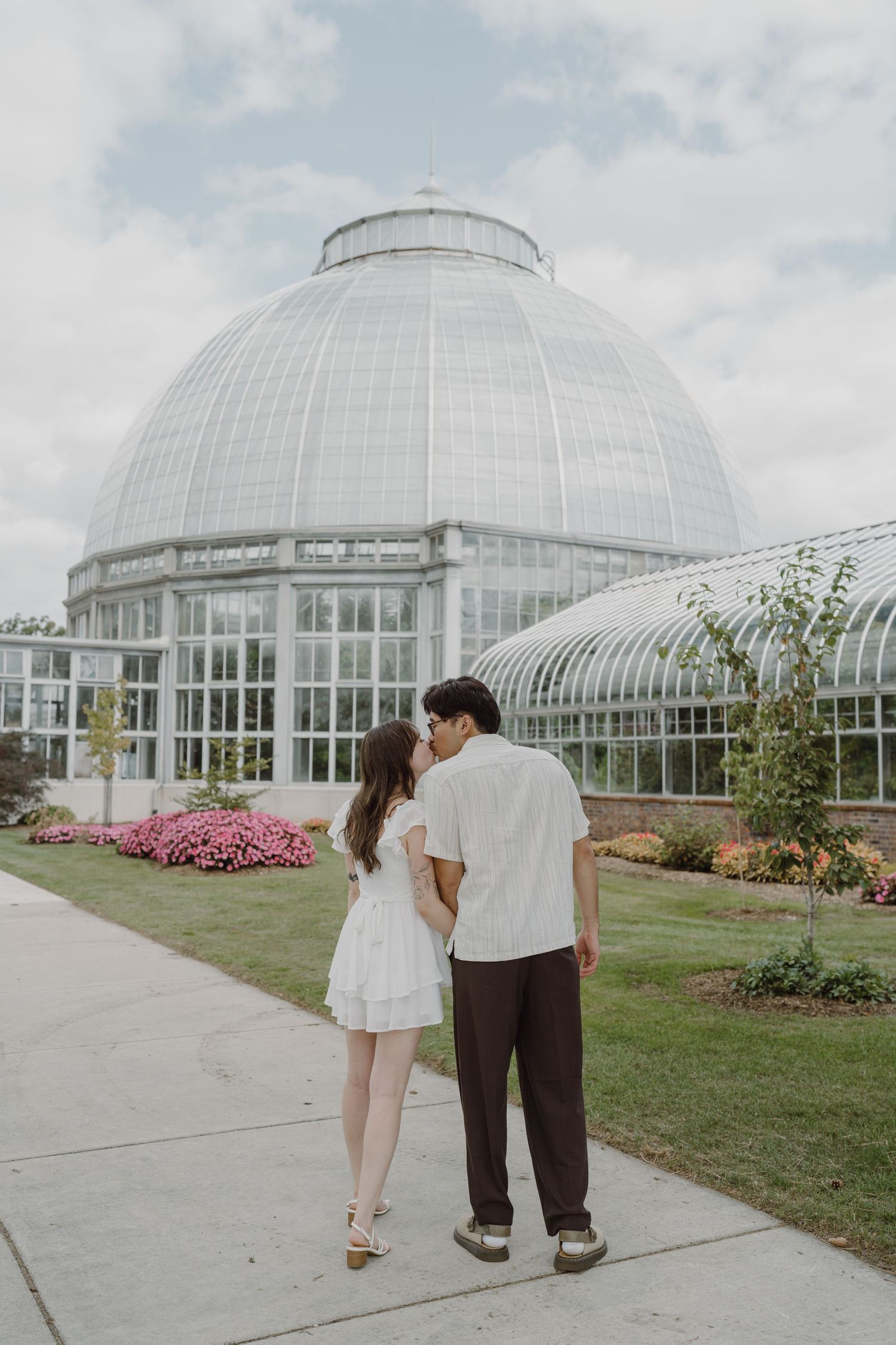 A couple walks hand in hand near a glass conservatory with landscaped gardens.