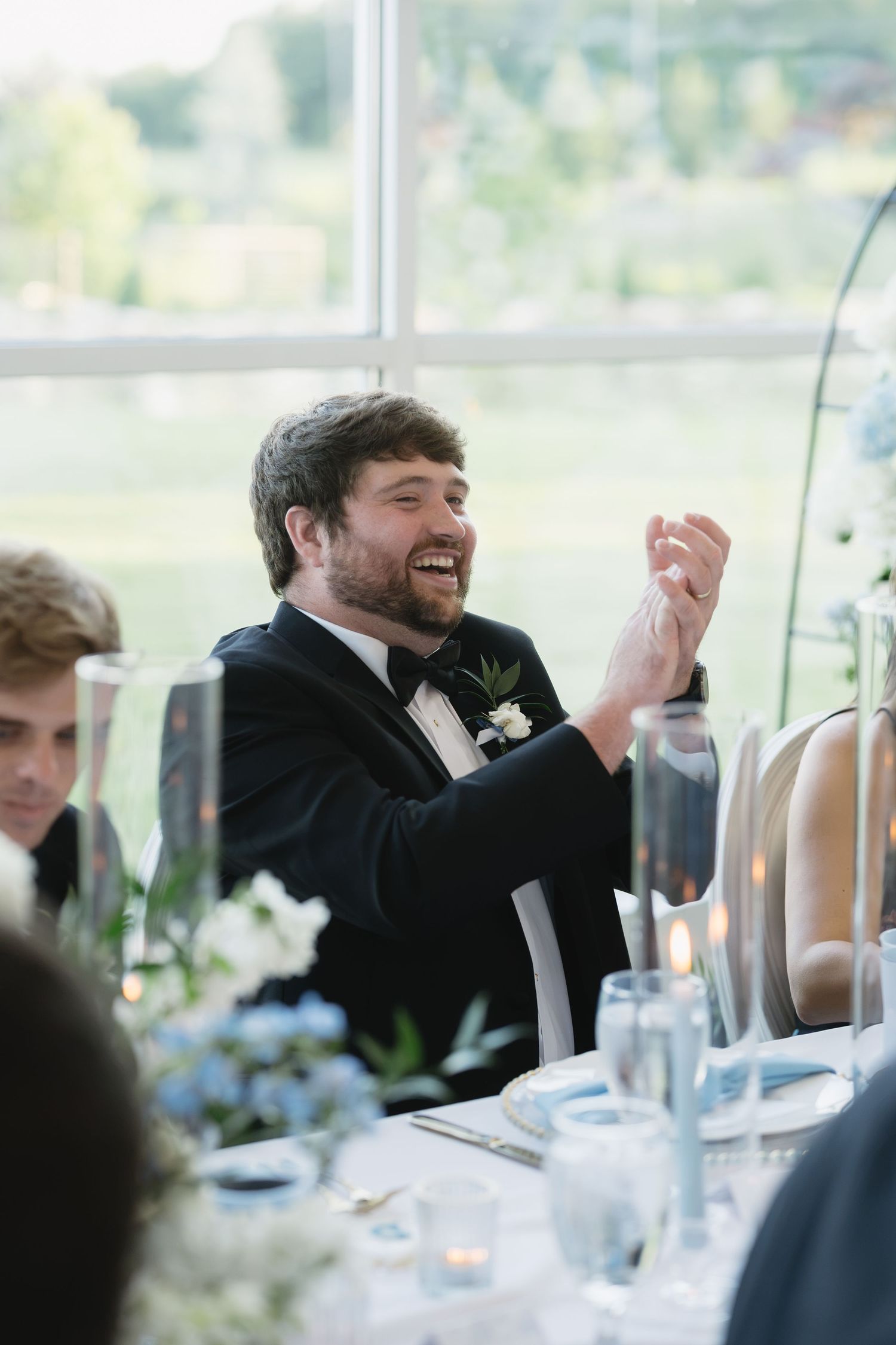 Groom in black tuxedo laughing joyfully at wedding reception table with blue floral centerpieces.