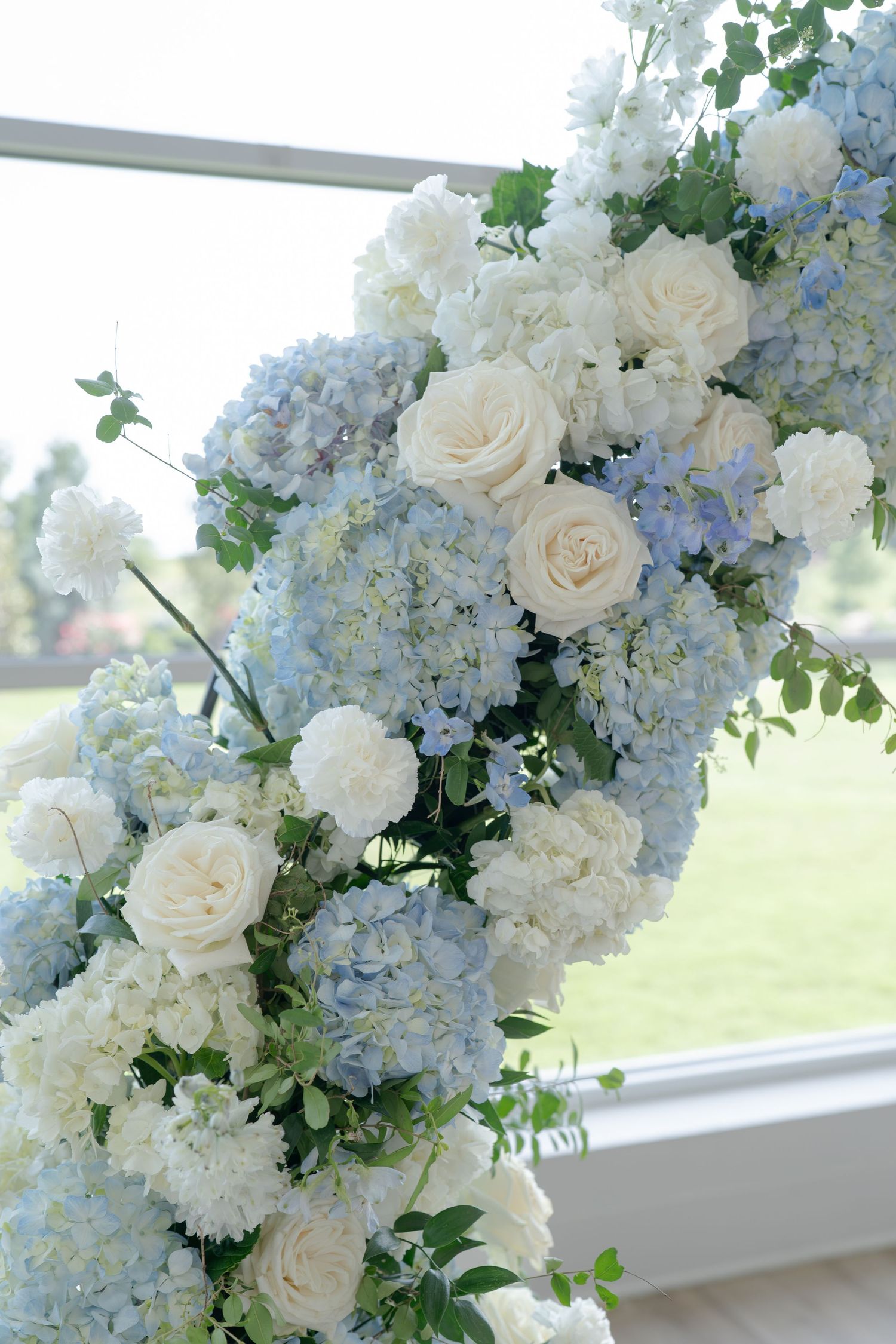 Elegant wedding arch adorned with soft blue hydrangeas and cream roses against a bright window backdrop.