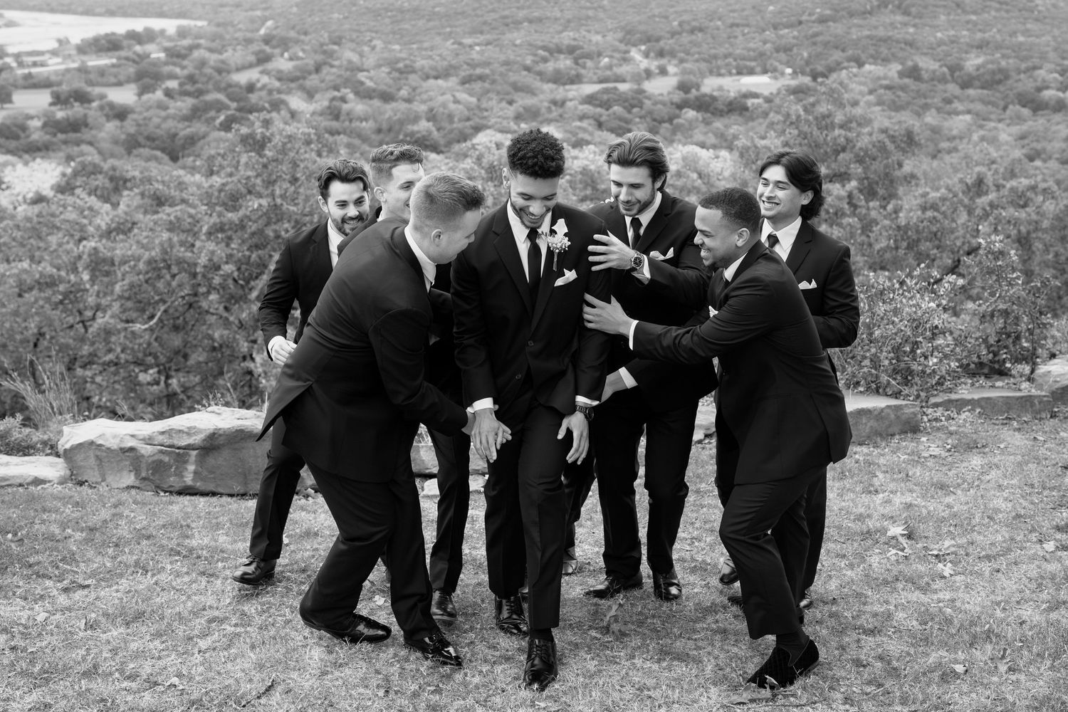 A group of groomsmen in black suits share a playful moment on a hilltop.