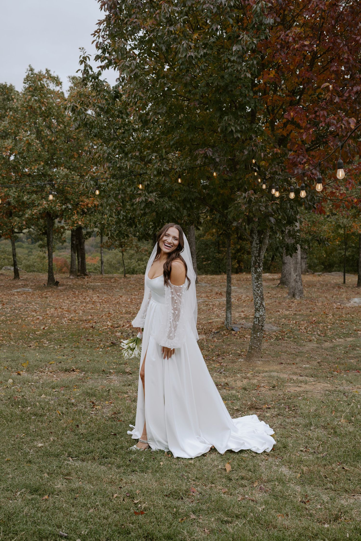A white wedding gown with detailed lace sleeves shown in a natural outdoor setting with string lights.