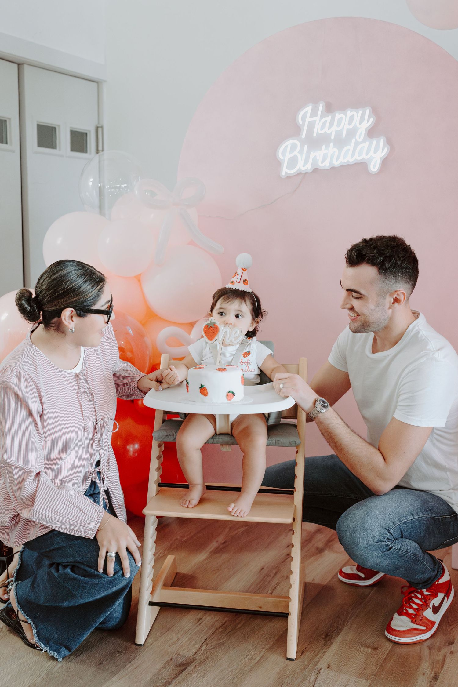 Birthday party setup with pink balloons and 'Happy Birthday' neon sign against a wall.