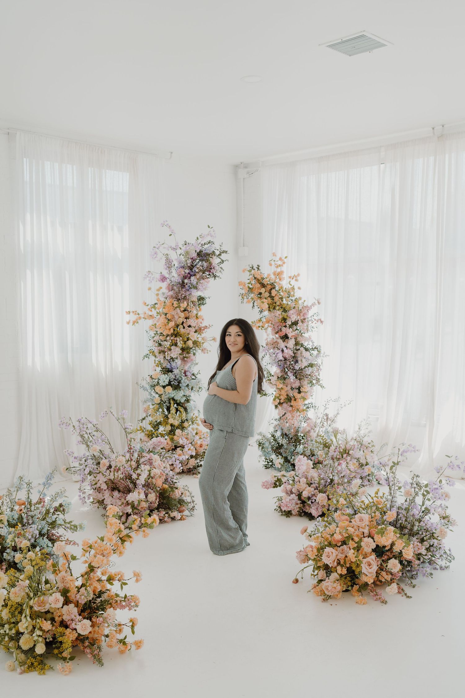 Maternity photo featuring soft floral arrangements and white curtains in minimalist studio.