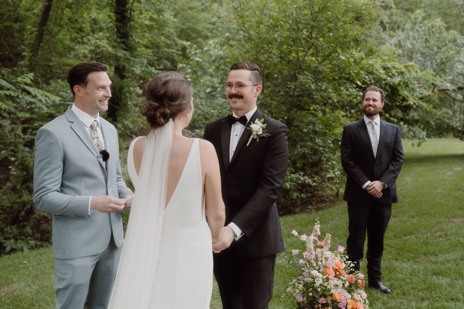 An outdoor wedding ceremony takes place in a lush green garden setting with colorful floral arrangements.