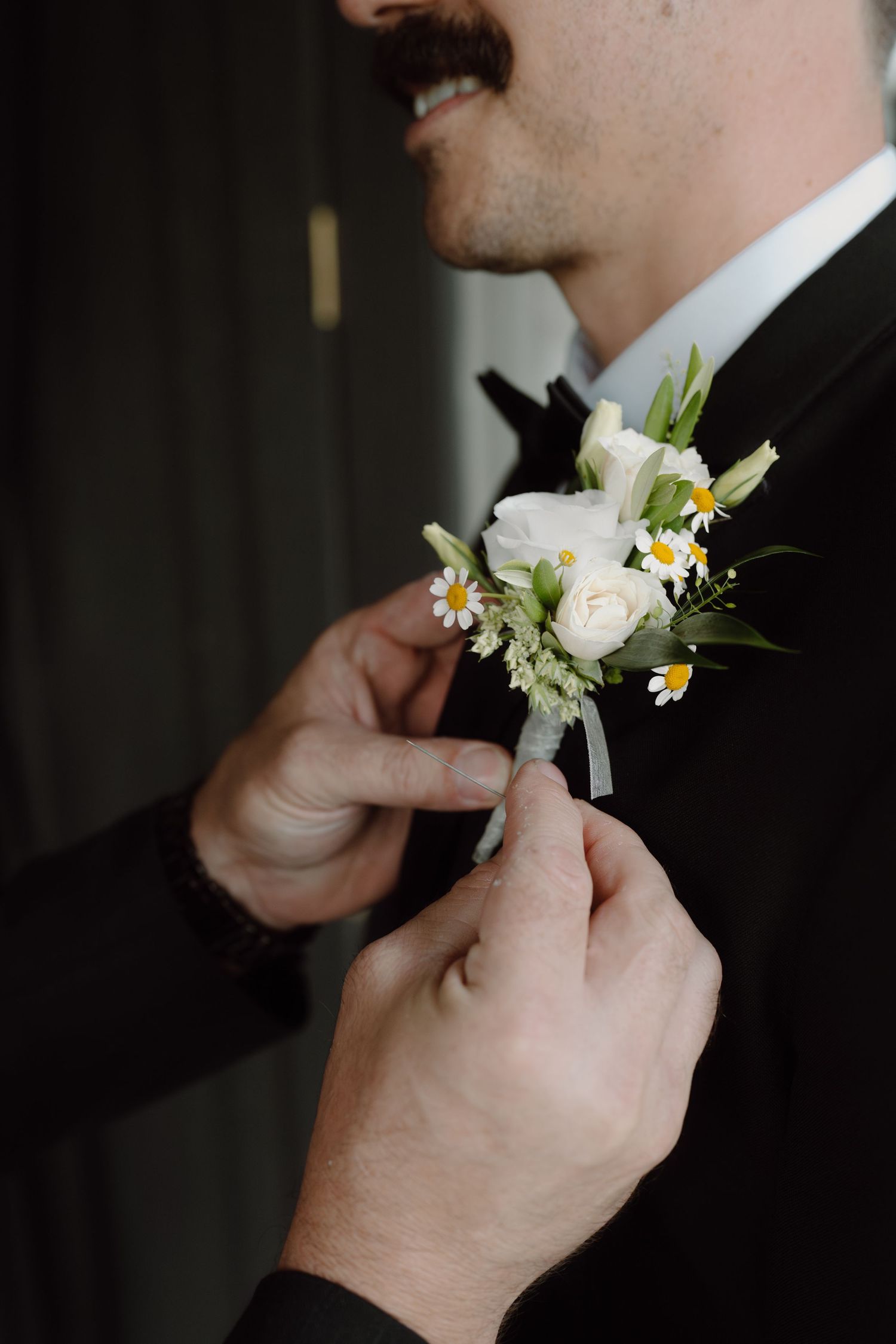 Close-up shots of hands pinning white floral boutonnieres onto formal suits.
