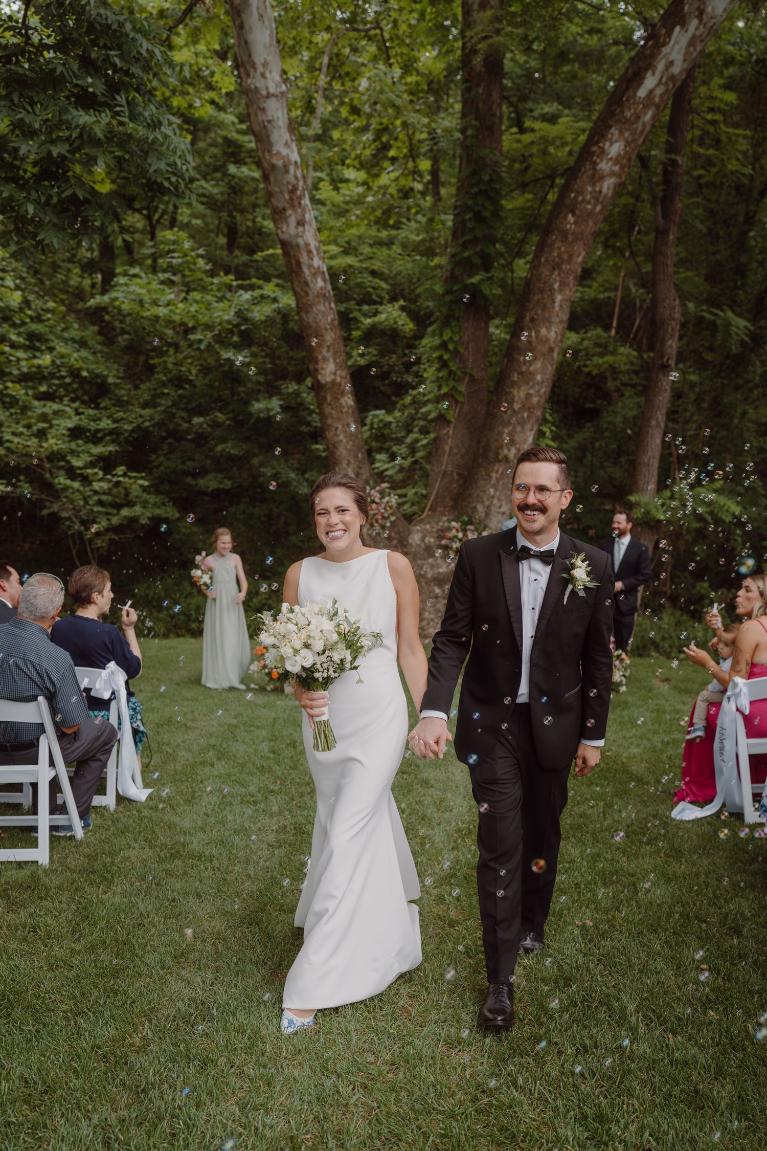 Newlyweds walk down a grassy aisle while guests celebrate in a forest setting.