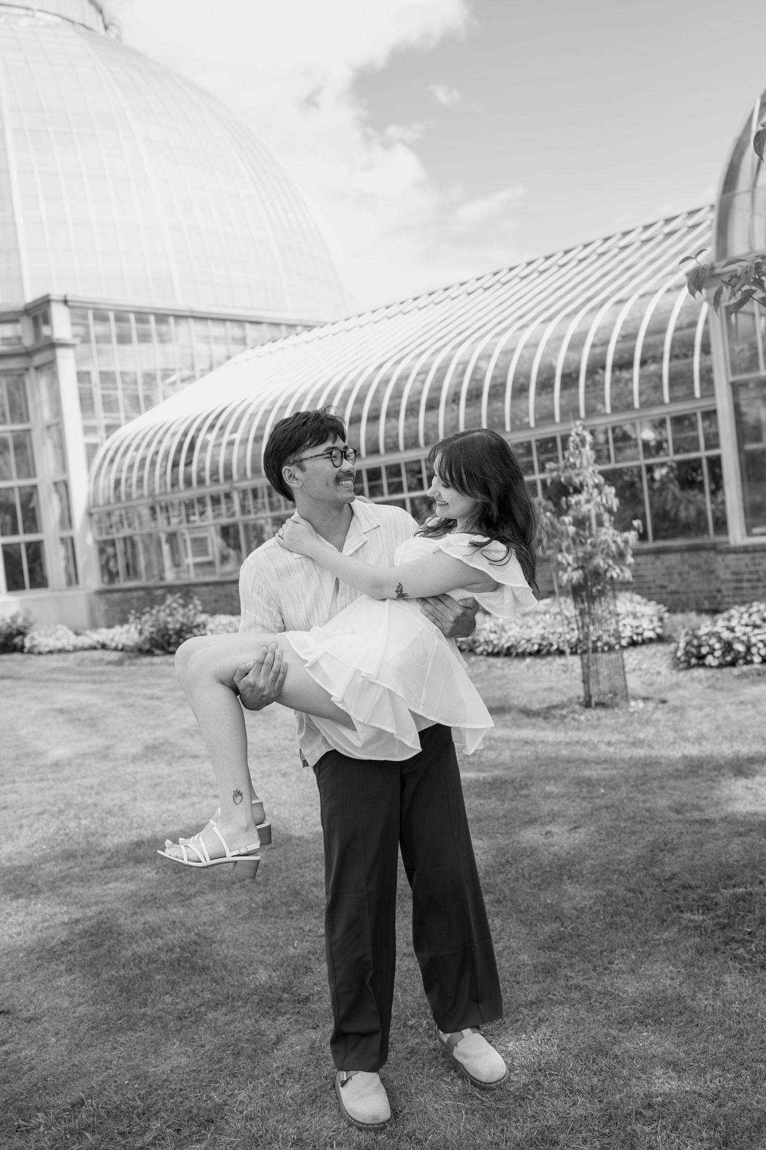 Black and white photo of a couple dancing outdoors with a greenhouse structure in the background.