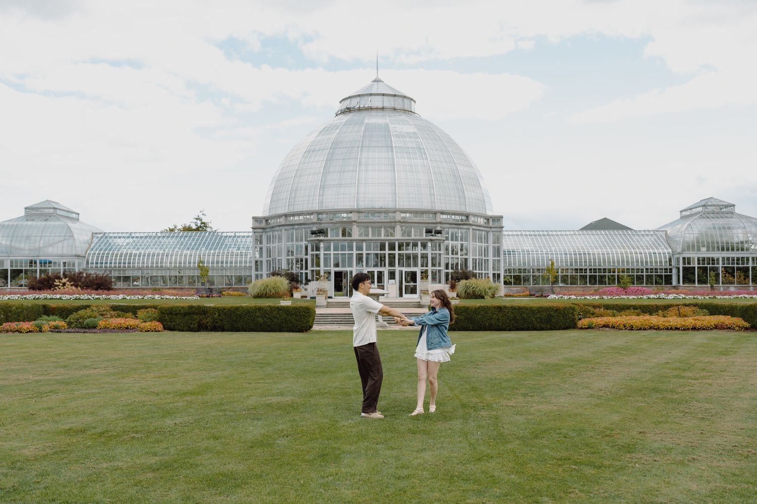 A couple poses in front of a large glass conservatory dome on a sunny day with green lawn in the foreground.