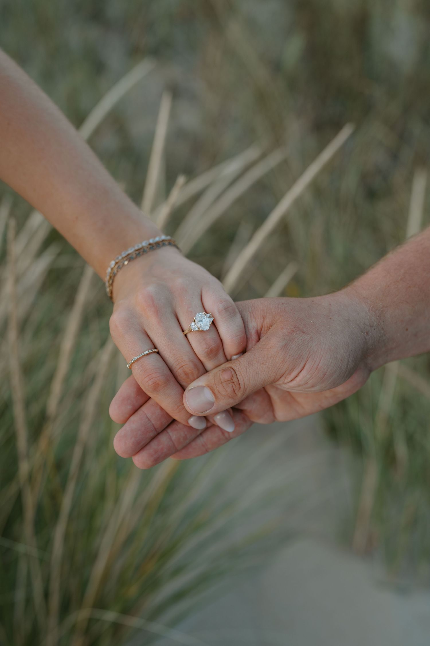 Intimate close-up of hands with engagement rings among coastal grass at sunset.
