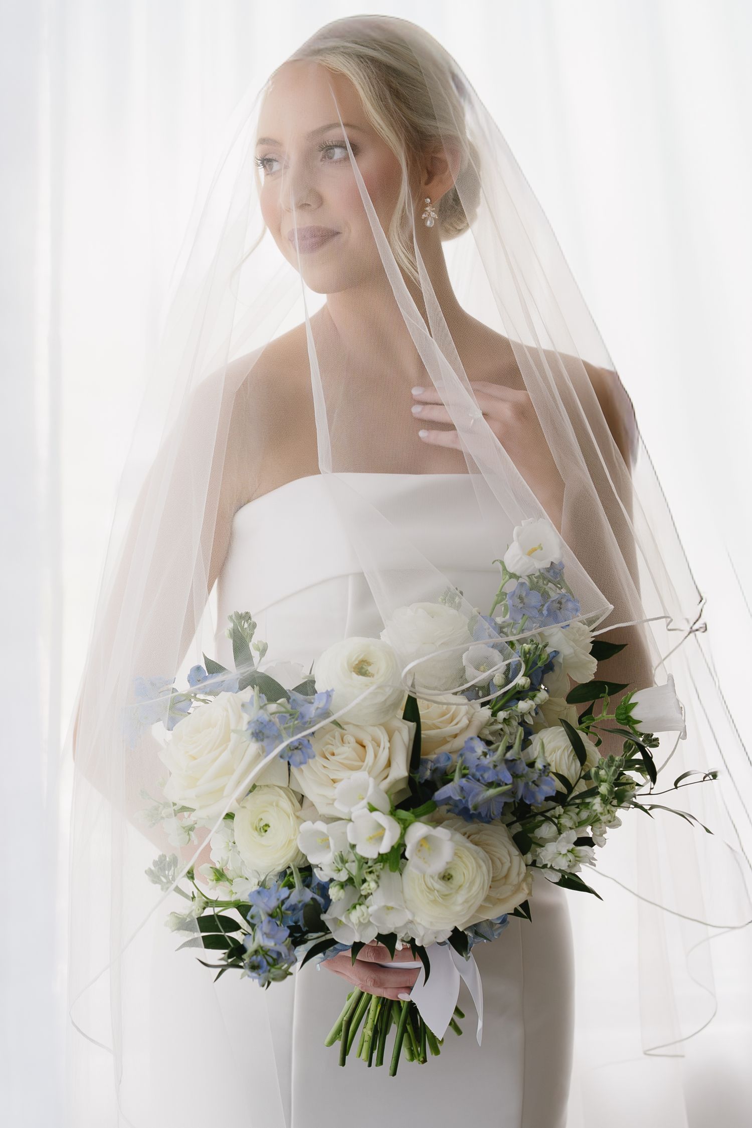 Bride in white strapless gown and veil holding blue and white floral bouquet with roses and delphinium.