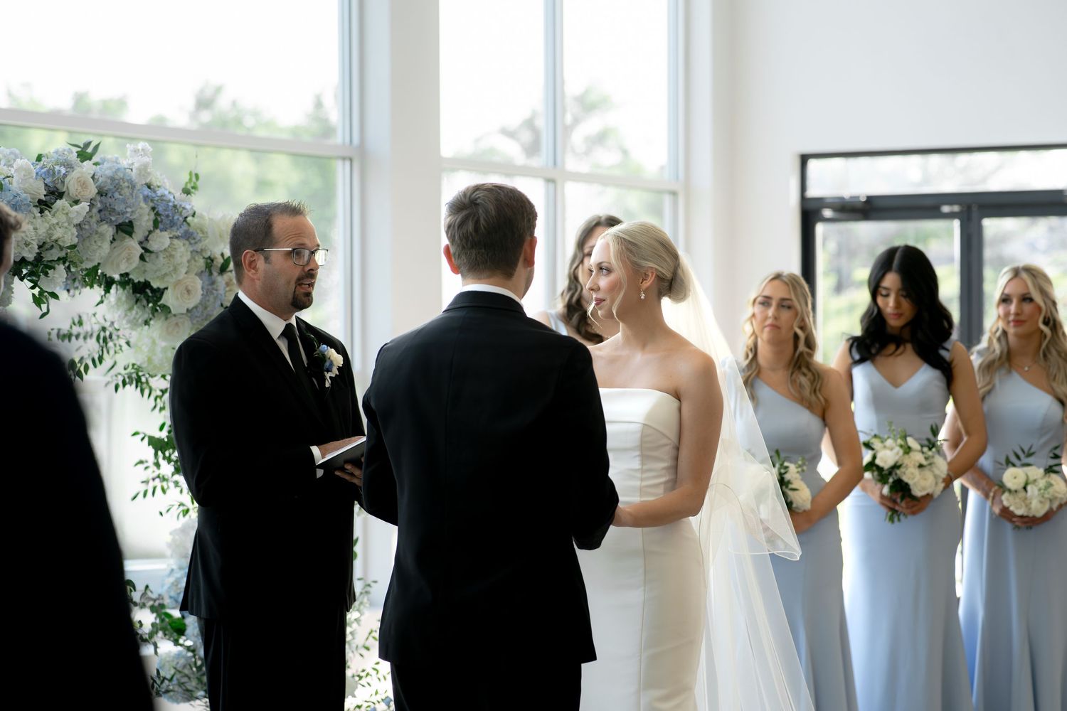 Bride and groom exchange vows at elegant indoor wedding ceremony with bridesmaids in light blue dresses.