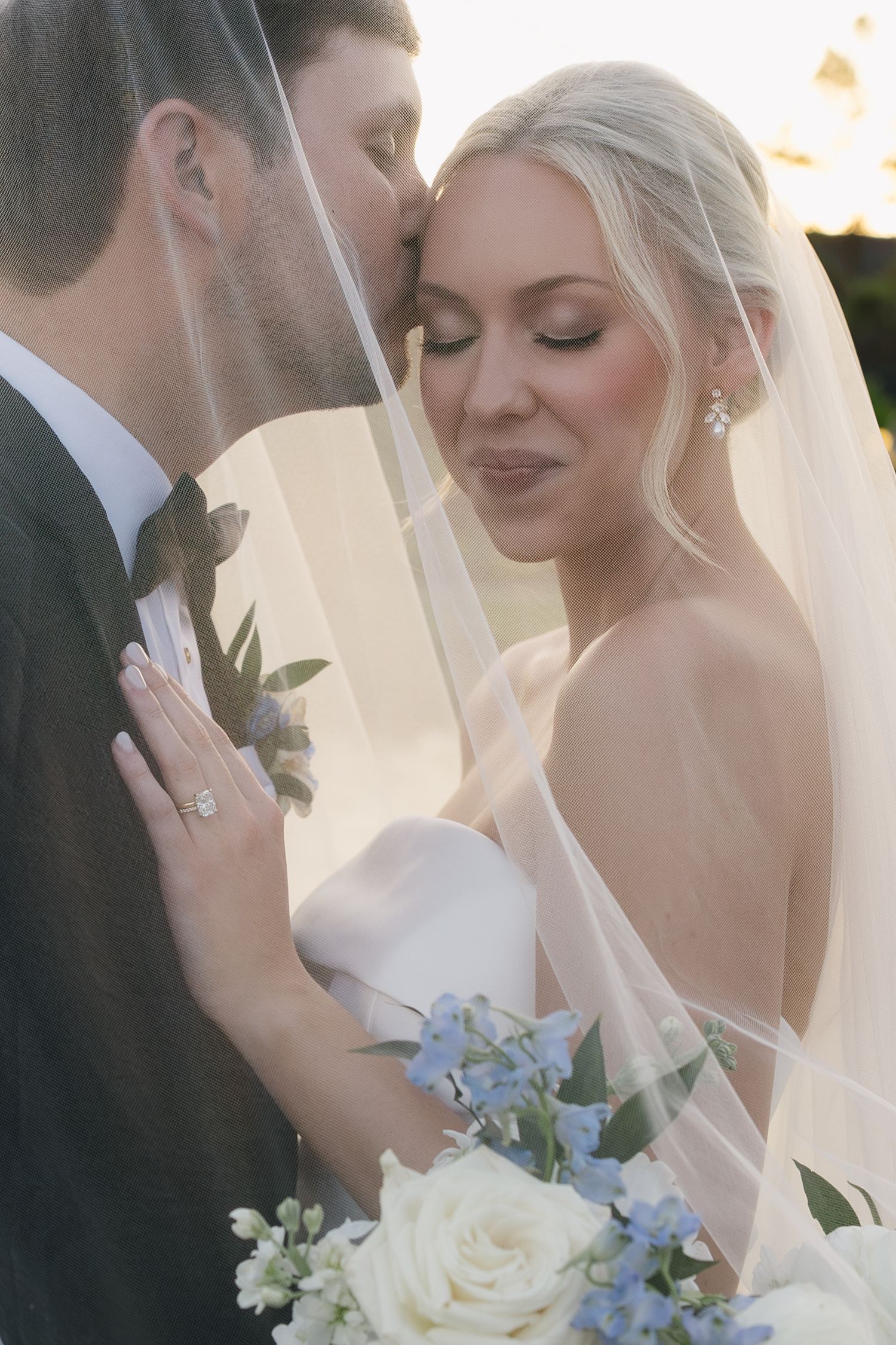 Bride and groom share a romantic moment under a sheer veil, bride holding white and blue floral bouquet.