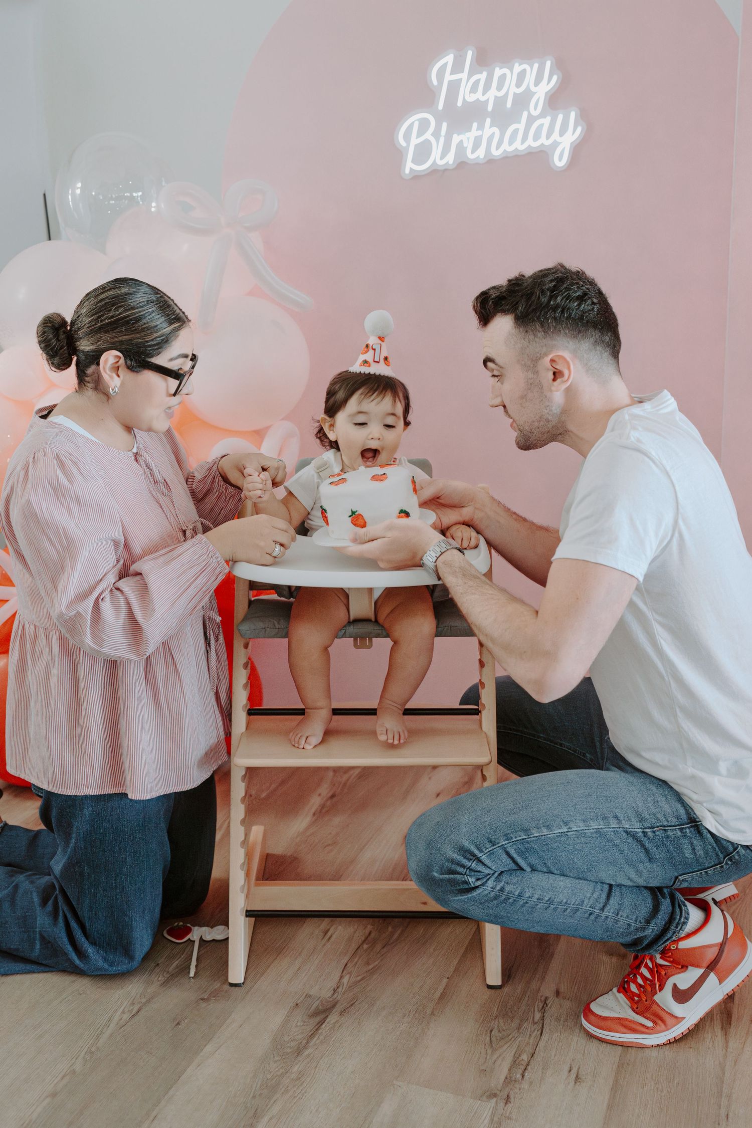 Family celebrating birthday with cake against pink backdrop and balloon decorations.