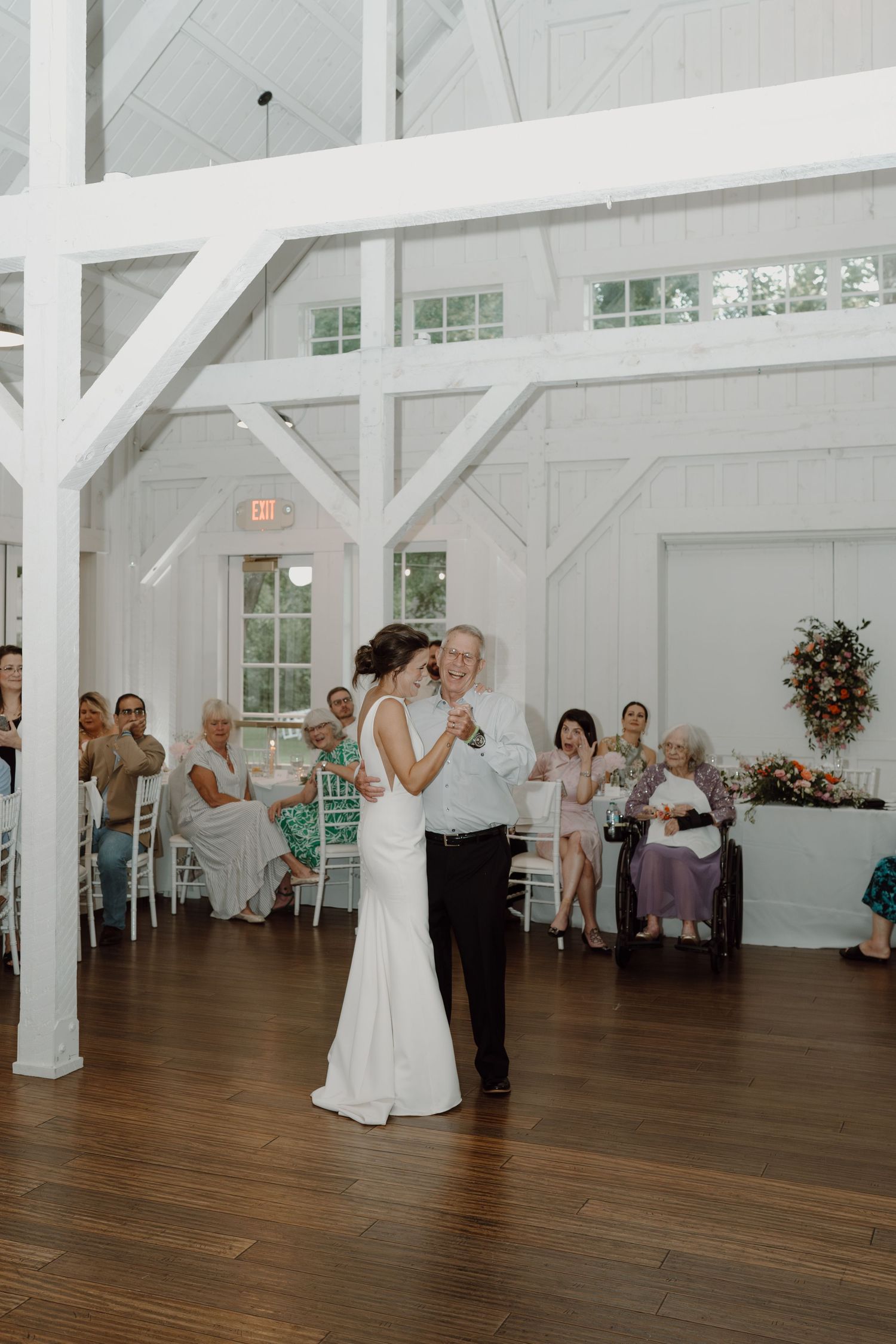 Dancers share a tender moment on the wooden floor of a bright white barn venue as guests look on.