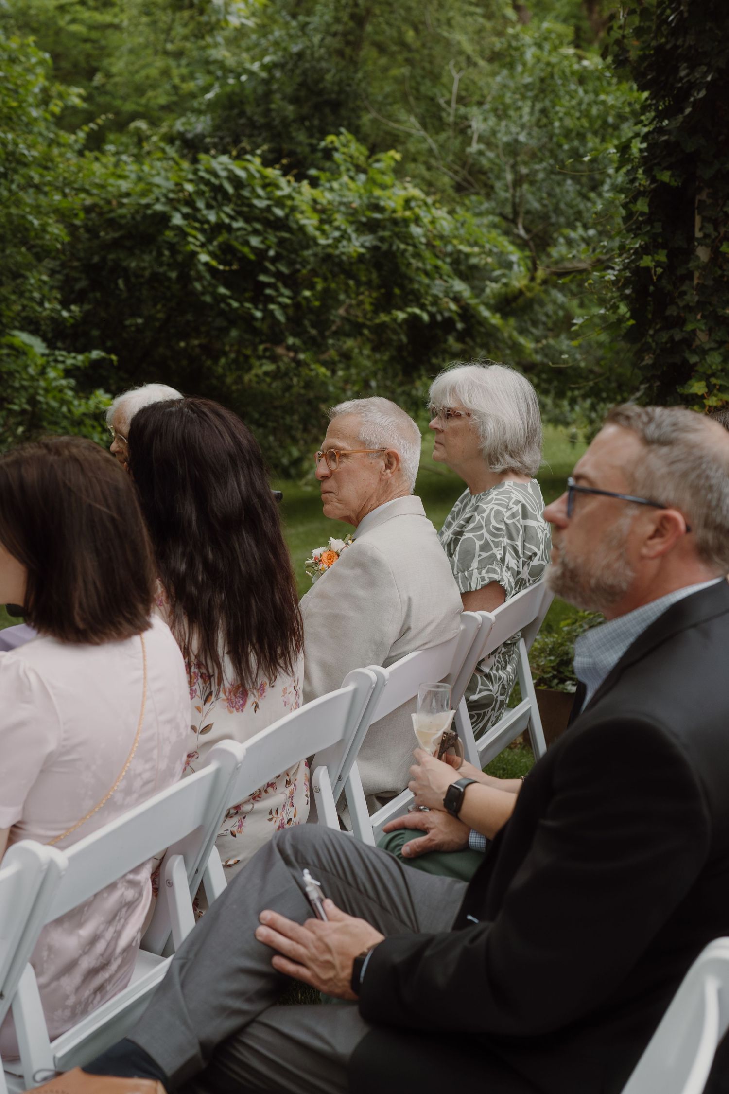 Wedding guests seated in white chairs outdoors watching ceremony in a wooded setting.