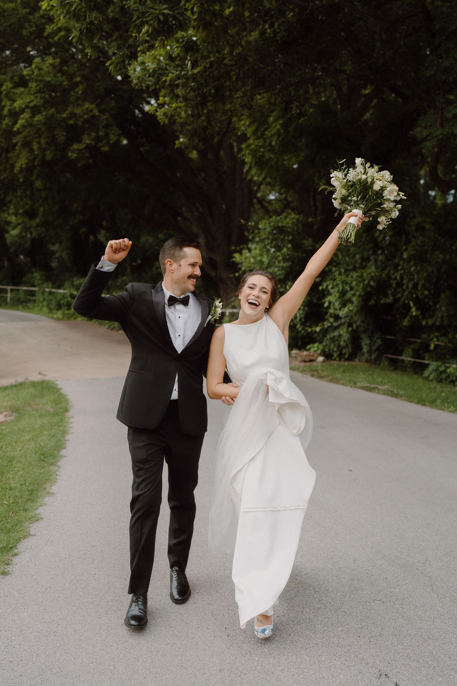 Happy couple celebrates with raised arms while walking down a scenic pathway on their wedding day.