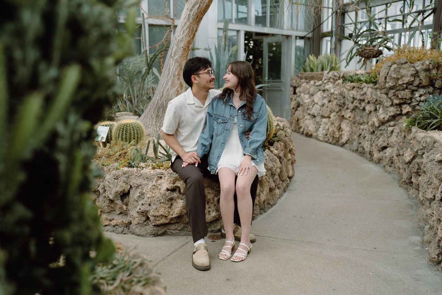 A couple relaxes on concrete steps surrounded by greenery and stone walls.