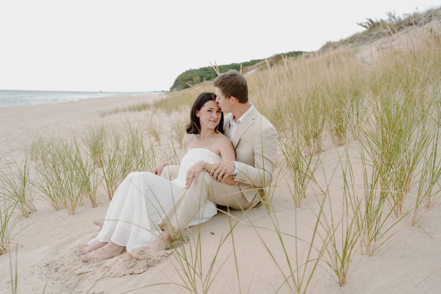 A couple embraces on a sandy beach with tall grass in the background during sunset.