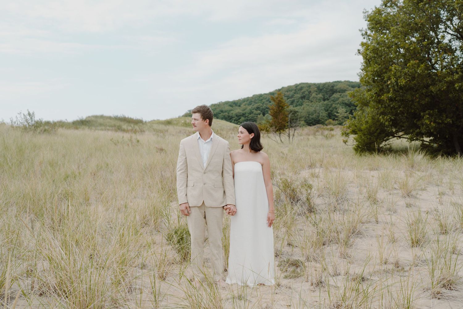 A couple in light colored formal wear poses together on sandy dunes with hills behind them.