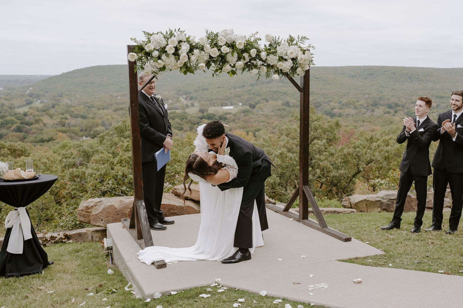 A romantic wedding ceremony takes place on a hilltop with a wooden arch adorned with white flowers and greenery.
