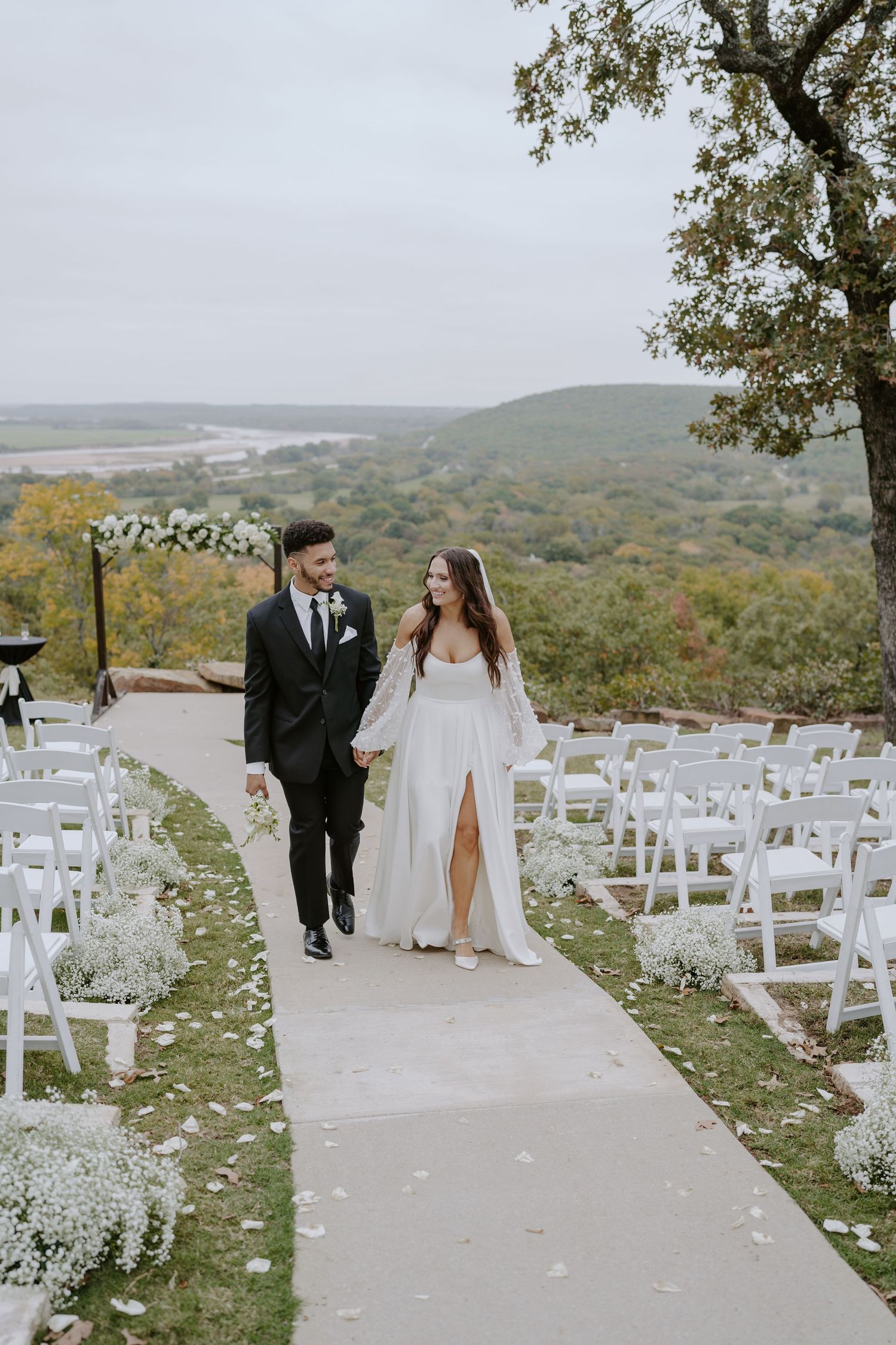 A hilltop wedding ceremony site with white chairs and floral arrangements against a mountain backdrop.