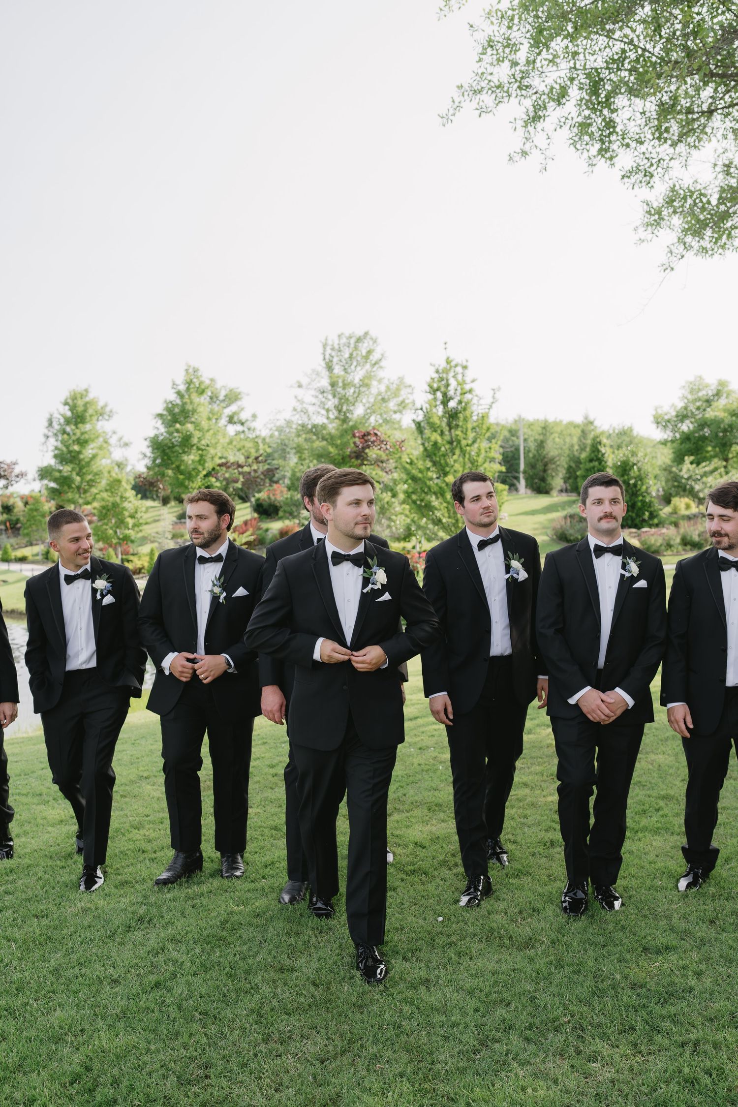 Groom and groomsmen in black tuxedos with bow ties walking outdoors on green grass at a wedding.
